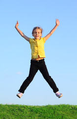 Happy little girl jumps at green grass at background of blue sky