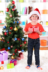 Little boy in Santa hat stands near Christmas tree with gift in