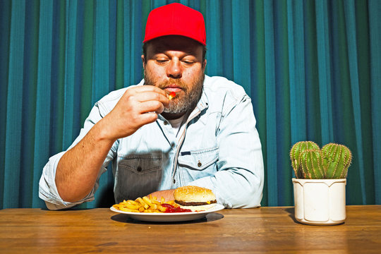 Man With Beard Eating Fast Food Meal. Trucker With Red Cap.