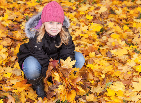 Little blond girl seat in the park with yellow leaves