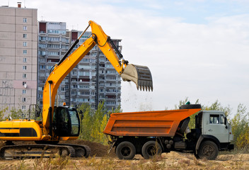 Excavator loads the ground in a truck on a background of houses