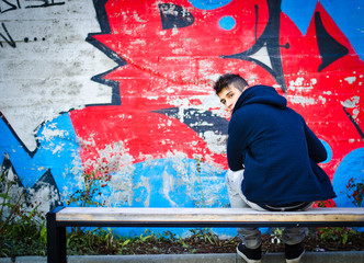 Young man with hoodie sitting on bench in front of graffiti