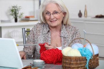 Senior woman knitting in front of her laptop computer
