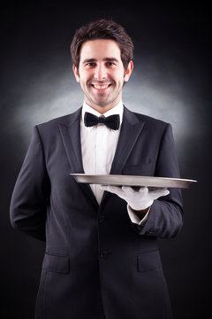 Portrait of a young waiter holding an empty dish on black backgr