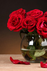 Beautiful red roses in vase on wooden table on brown background