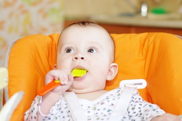 The baby with a spoon sits on a babies chair