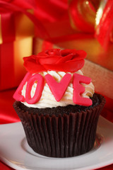 Cupcake with a red rose on top and gifts in boxes on red satin b