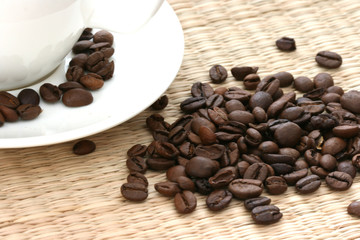 Whole coffee beans scattered on straw background with white cup