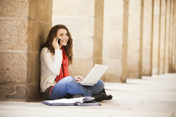Caucasian woman sitting on ground with laptop and talking on cell phone