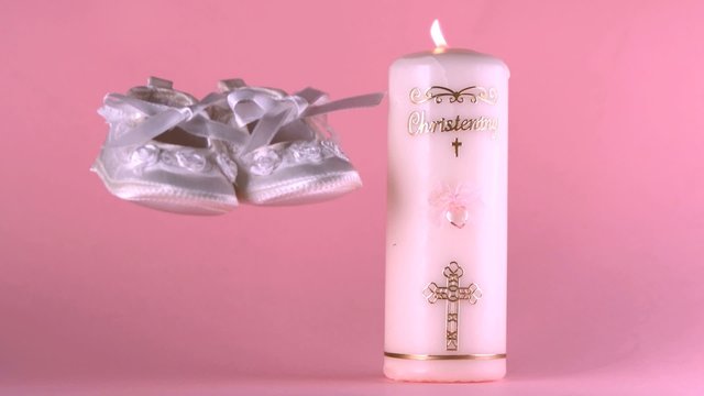 Baby shoes falling beside lit baptism candle on pink background