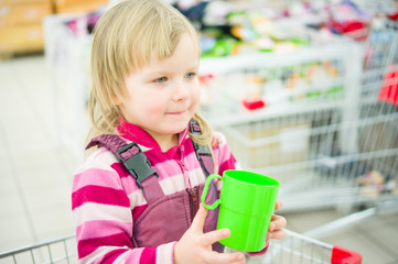 Adorable girl on shopping cart with green cup in supermarket