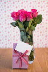 Bunch of pink roses in vase with pink gift leaning against it an