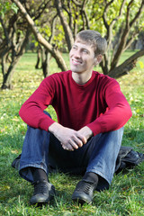 Naklejka na ściany i meble Young man in red sweater smiles and sits on grass in park