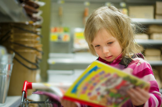 Adorable girl in shopping cart select books on shelves in superm