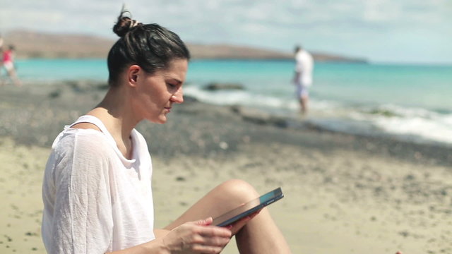 Woman sitting on the beach with tablet, family in background