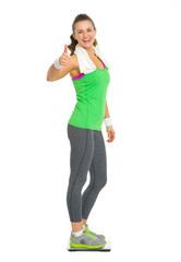 Smiling fitness woman standing on scales and showing thumb up