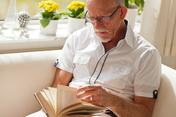 Senior man with glasses reading book in living room.