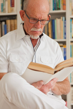 Senior man with glasses reading book in front of bookcase.
