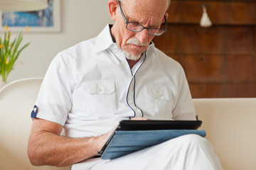 Senior man with glasses using tablet on couch in living room.