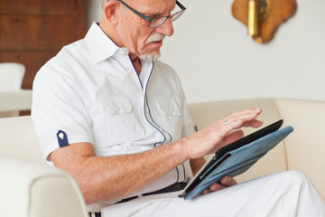 Senior man with glasses using tablet on couch in living room.