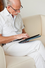 Senior man with glasses using tablet on couch in living room.