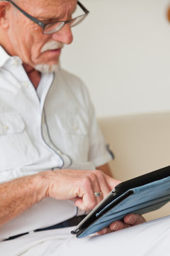 Senior man with glasses using tablet on couch in living room.