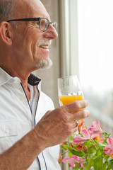 Senior man with glasses drinking orange juice at window with flo