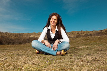 Smiling pretty brunette woman wearing jeans sitting in meadow wi