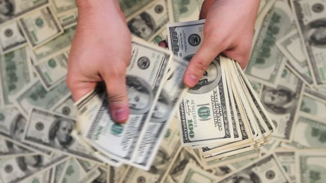 Looking down at hands counting a stack of hundred dollar bills with money in the background.
