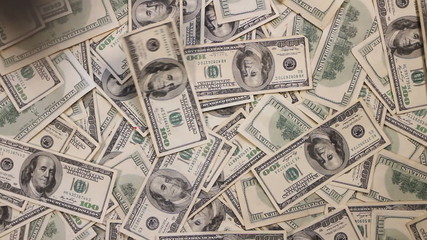 Looking down at paper bills falling one by one into a pile of hundred dollar bills. 