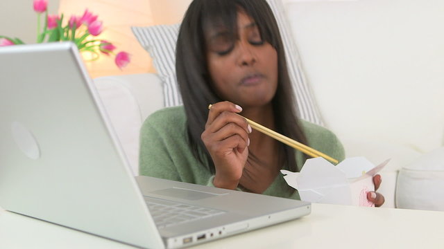 Black woman eating Chinese take out while using laptop