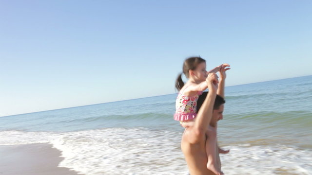 Father Running Along Beach Carrying Daughter On Shoulders