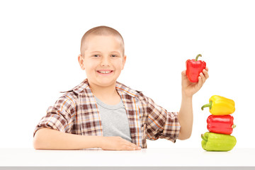 A little smiling boy holding colorful peppers on a table