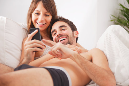  Smiling young couple looking smartphone in bed