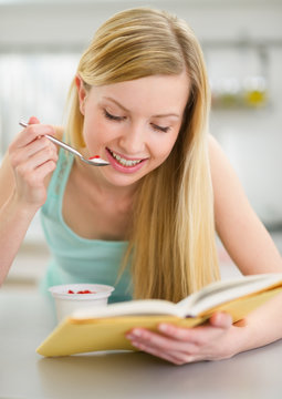 Happy teenager girl reading book and eating yogurt in kitchen