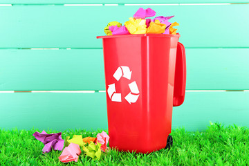 Recycling bin with papers on grass on light blue background