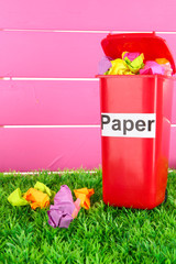 Recycling bin with papers on grass on pink background