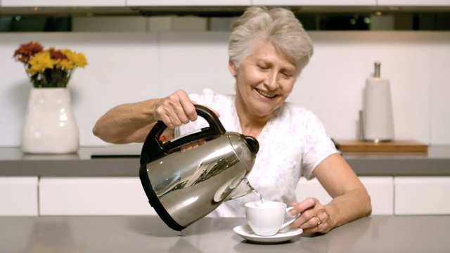 Elderly Woman Pouring Boiling Water From Kettle Into Cup