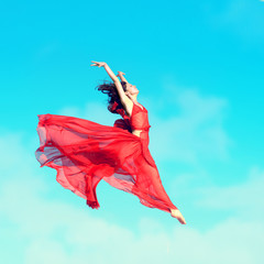 Woman in airy red dress jumping in the air, blue sky background