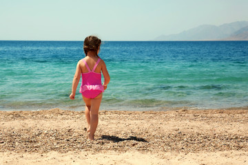 Fototapeta na wymiar Little girl standing on beach in swimsuit and going to swim