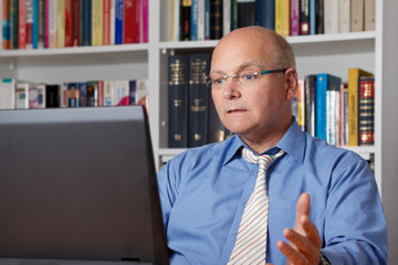 Frustrierter Geschäftsmann sitzt vor Computer oder Laptop, Bücher im Hintergrund.