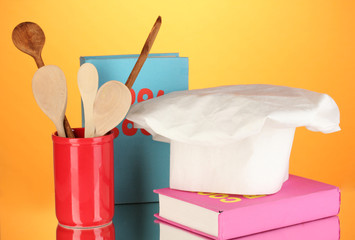 Chef's hat with spoons and cook book on orange background