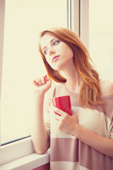 Beautiful young woman with cup over window.