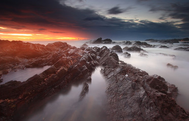 Dramatic sunrise over rocky shoreline with smoky sea