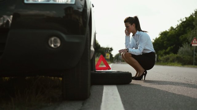 Young woman with flat car tyre calling tow truck by phone