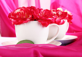 Roses in cups on wooden table on pink cloth background