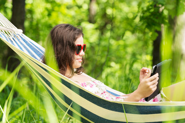 Young woman lying in a hammock in garden with E-Book.