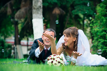 groom and bride lie on a grass around soap bubbles