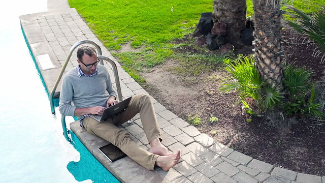 Young man working with laptop while sitting by swimming-pool, vi