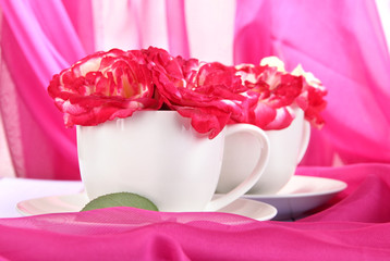 Roses in cups on wooden table on pink cloth background